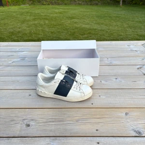 Valentino Garavani vita sneakers med mörk blå detalj - Säljer ett par vita Valentino Garavani sneakers med bred mörk blå rem över mitten och diskret logga på sidan. Skorna har rund tå, platt sula och är tillverkade i skinn. Klassisk och stilren design med box och dustbag medföljer. Enligt denna förra ägaren så är dessa äkta men vet inte! Skriv om ni har funderingar eller om ni vill ha fler bilder!