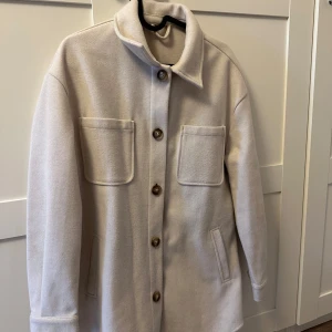 Skjortjacka Beige - Säljer en stilren beige overshirt jacka med stora bröstfickor och knappar framtill. Jackan har klassisk krage och raka ärmar, perfekt för lager-på-lager. Materialet känns mjukt och har en ull-liknande finish.