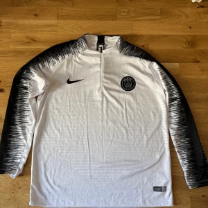 PSG långärmad träningströja Nike M - Snygg långärmad Paris Saint-Germain träningströja från Nike i vitt och svart. Tröjan har halv dragkedja, svart Nike-logga och PSG-märke på bröstet. Ärmarna är svarta med vitt grafiskt mönster. Tillverkad i lätt och ventilerande material. Aldrig använd