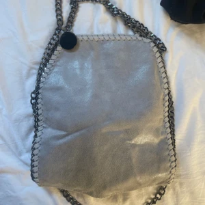 Grå axelväska med kedjedetaljer - Snygg grå axelväska med metallic finish och kraftiga kedjor som axelrem. Väskan har dekorativa sömmar längs kanterna och en rund metallbricka vid remmen. Perfekt för dig som gillar en edgy och modern stil.