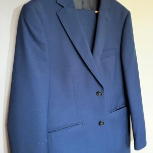 Mörkblå kavaj från Dressmann med byxor! - Snygg mörkblå kavaj från Dressmann med klassisk passform. Kavajen har två knappar framtill, tre fickor och är helfodrad med svart och ljusblått foder. Perfekt för dig som vill ha en stilren och tidlös look. I väldigt bra skick ✨