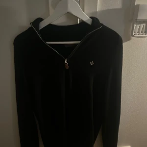 Svart merinoullströja från Morris - Svart långärmad tröja från Morris i mjuk merinoull. Tröjan har en stilren half zip-dragkedja och en liten broderad logga på bröstet. Perfekt för dig som gillar klassisk och clean stil med en modern touch. PRIS KAN DISKUTERAS 