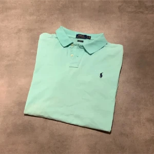 Ljusblå pikétröja från Polo Ralph Lauren - Säljer en klassisk ljusblå pikétröja från Polo Ralph Lauren med broderad logga på bröstet. Tröjan har korta ärmar, krage och knäppning framtill. Perfekt för en stilren och avslappnad look. Tröjan är blekt som ni ser på de vita prickarna och har någon röd färg som ni också ser på en av bilderna. Pris kan diskuteras vid snabb affär! Bara att höra av sig vid funderingar eller frågor!😀
