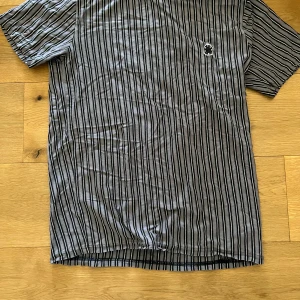 Randig t-shirt från Woodbird - Säljer en randig t-shirt från Woodbird i svart och grått. T-shirten har korta ärmar och en liten broderad detalj på bröstet. Perfekt för dig som gillar en stilren och avslappnad look.