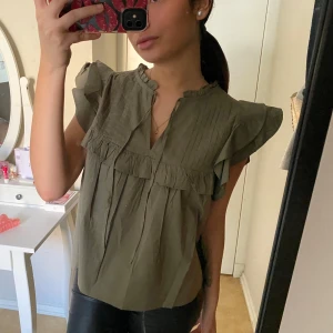Olivgrön blus med volangdetaljer - Säljer en olivgrön blus med volangdetaljer och korta ärmar. Blusen har en lätt v-ringning med liten krage och dekorativa sömmar framtill. Perfekt att styla med jeans eller kjol.