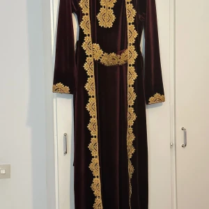 Mörk vinröd långklänning med broderade detaljer - Säljer en elegant mörk vinröd långklänning med vackra guldiga broderade detaljer längs hela framsidan, ärmsluten och midjan. Klänningen har lång ärm och är tillverkad i ett mjukt, lyxigt material som ger en exklusiv känsla.