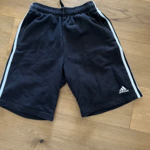 Svarta shorts från Adidas med vita ränder - Svarta Adidas shorts med vita ränder längs sidorna och klassisk logga på ena benet. Resår i midjan för skön passform. Perfekta för träning eller avslappnade dagar.