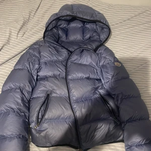 Blå Moncler pufferjacka med huva - Säljer en blå pufferjacka från Moncler med huva och dragkedjor på fickorna. Jackan har Moncler-logga på ärmen och är riktigt fluffig och varm. Perfekt för kalla dagar och har en snygg, sportig vibe. Passar dig som vill ha både stil och funktion. Hyfsat gammal modell som jag köpte från vestaire. Som ni kan se på bilderna så finns det hål som är igentäppta med en speciell ljusblå tejp. Hålen är inte märkvärdiga eftersom de ligger på underarmarna. Storlek small. 