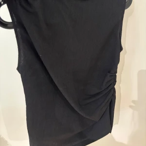 Svart ärmlös topp med rynk - Snygg svart ärmlös topp med rynkade detaljer på sidan som ger en cool och trendig look. Toppen har en enkel och stilren design, perfekt för dig som gillar minimalistiskt mode. Materialet känns lätt och följsamt.