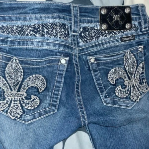 Miss Me blå bootcut jeans - Snygga blå jeans från Miss Me i bootcut-modell med coola paljettbroderier och stora dekorativa detaljer på bakfickorna. Jeansen har kontrastsömmar, blingiga knappar och är perfekta för dig som gillar statement-plagg det ända är att de har en liten grön fläck typ vid knät.