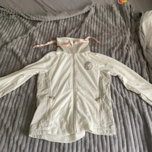 Vit fleecejacka från Jacson - Säljer en vit fleecejacka från Jacson med dragkedja och huva med ljusrosa snören. Jackan har långa ärmar, två fickor med dragkedja och ett broderat märke på bröstet. Perfekt för ridning eller som mysig vardagsjacka.