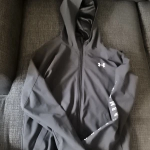 Svart vindjacka från Under Armour XS - Svart vindjacka från Under Armour med huva och hel dragkedja. Jackan har logga på bröstet och stort tryck längs ena ärmen. Lätt material och loose fit, perfekt för aktiva dagar. Snygg och sportig stil som funkar året runt.