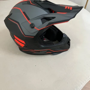 SHOT crosshjälm svart/röd - Snygg crosshjälm från SHOT i mattsvart och grått med röda detaljer. Hjälmen har en sportig, kantig design med skärm framtill och ventiler för bra luftflöde. Invändigt finns mjuk, vadderad stoppning för extra komfort. Perfekt för motocross eller enduro. E märkt 