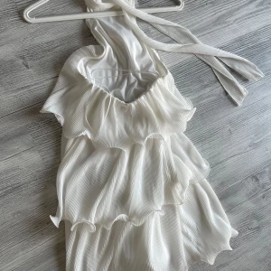 Studentklänning Gina Tricot - Superfin vit kortklänning från Gina Tricot med plisserat tyg och flera volanger. Axelbandslös men med ett knyte runt halsen!