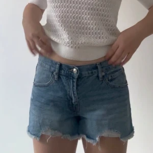Blå jeansshorts med fransig kant - Snygga blå jeansshorts med fransig rå kant och klassisk femficksdesign. Shortsen har normal passform och midwaist, perfekt för en chill sommarlook. De är tillverkade i jeansmaterial och har knapp- och dragkedjestängning framtill.