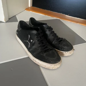 Svarta sneakers från Axel Arigato - Svarta sneakers från Axel Arigato med vit sula och snörning. Skorna har en stilren design med fågelbroderi på sidan och textdetalj '1.40MM' på hälen. Tillverkade i skinn och passar perfekt till en urban streetstyle.