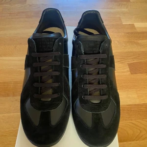 Maison Margiela - Säljer ett par svarta sneakers från Maison Margiela med snygga detaljer i mocka och skinn. Skorna har rund tå, platt sula och klassisk snörning. Perfekta för dig som gillar stilrena och exklusiva sneakers.
