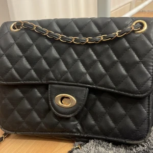 Svart quiltad axelväska med guldkedja - Säljer en svart quiltad axelväska med guldfärgade detaljer och kedja. Väskan har ett klassiskt rutmönster, vridlås framtill och innerfack med dragkedja. Perfekt för dig som vill ha en stilren och tidlös accessoar.