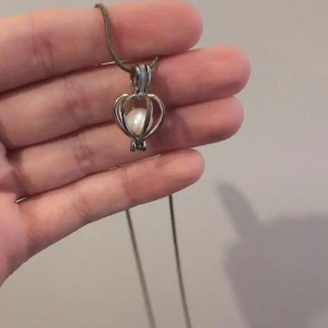 Halsband med pärlhänge i silverfärg - Säljer ett halsband med ett unikt hänge i silverfärg, format som ett hjärta med en vit pärla i mitten. Kedjan är tunn och enkel, perfekt för att ge en elegant touch till din stil. Lite rostig men inte märkbart långt ifrån eller när den rör sig.