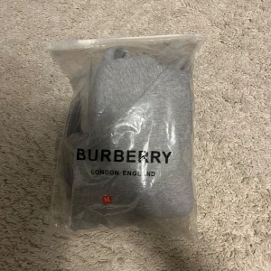 Grå hoodie från Burberry - Säljer en grå hoodie från Burberry med klassiskt rutigt foder i huvan. Tröjan har dragkedja framtill, justerbar huva med snören och ribbade muddar. Perfekt för dig som gillar stilrena och exklusiva plagg.