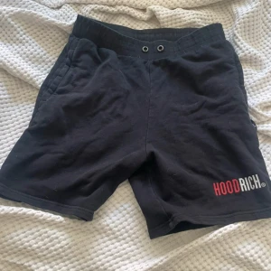 Svarta shorts från Hoodrich - Svarta shorts från Hoodrich med tryckt logga i rött och vitt på benet. Modellen har elastisk midja med två metallöljetter framtill och sidofickor. Perfekta för chill eller streetwear. Mjukt material som känns skönt mot huden.