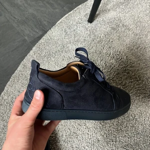 Mörkblå sneakers från Louboutin - Snygga mörkblå sneakers från Louboutin i mocka med klassisk röd sula och broderad logga på hälen. Skorna har rund tå, platt sula och snörning. Perfekta för dig som vill sticka ut med exklusiva detaljer och lyxig känsla. Saknar box.
