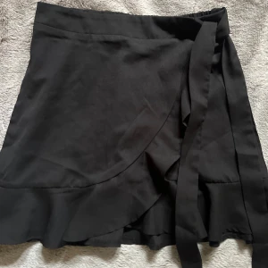 Svart omlottkjol med volang från SHEIN - Svart kort omlottkjol från SHEIN i storlek XS. Kjolen har volangkant nertill och knytband i midjan för en snygg passform. Perfekt för dig som gillar en enkel men trendig look.