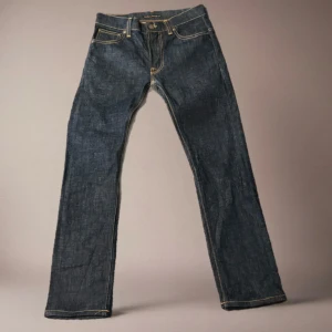 Mörkblå jeans från Nudie Jeans - Snygga mörkblå jeans från Nudie Jeans med klassisk femficksdesign och kontrastsömmar. Byxorna har raka ben och ikoniska broderade bakfickor. Tillverkade i slitstarkt jeansmaterial av bomull. Perfekta för dig som gillar stilrena och tidlösa jeans.