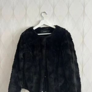 Svart pälsjacka  - Mysig svart pälsjacka från Vero Moda med rund halsringning och långa ärmar. Jackan har en fluffig känsla och stängs framtill med knappar. Normal i storlek, fint skick! 
