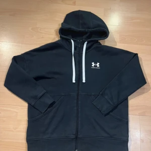 Svart hoodie från Under Armour - Svart hoodie från Under Armour med vit logga på bröstet och vita snören i huvan. Tröjan har dragkedja framtill, två stora fickor och är fodrad med mjuk fleece på insidan. Perfekt för chill eller träning, klassisk och enkel stil.