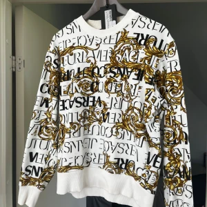 Vit Versace Tröja (M kan vara bra på S) - Säljer en vit sweatshirt från Versace Jeans Couture med svart logotypprint och barockinspirerade detaljer i guld över hela tröjan. Lyxig stil och använd 20 ish gånger.