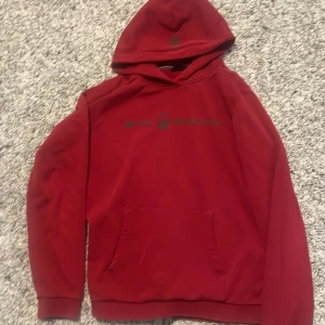 Röd hoodie från Sail Racing - Röd hoodie från Sail Racing med svart logga på bröstet. Klassisk känguruficka framtill och huva. Perfekt för dig som gillar en sportig och avslappnad stil. Mjuk insida och ribbade muddar vid ärmslut och nederkant.