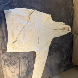 Grå Essentials Fear of God hoodie - Säljer en ljusgrå hoodie från Fear of God Essentials med tryck på bröstet och stor text på ryggen. Tröjan har huva, känguruficka och långa ärmar med ribbade muddar. Perfekt för dig som gillar streetwear och clean stil.