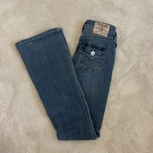 True Religion Becca Bootcut Jeans - Säljer ett par snygga blå bootcut jeans från True Religion, modell Becca Mid Rise Bootcut. Jeansen har klassisk femficksdesign, coola detaljer på bakfickan och en midwaist passform. Materialet är mjukt jeans och färgen är ljusblå med lätt tvättad look.
