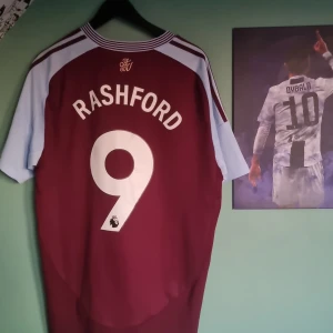 Aston Villa Rashford äkta helt ny - Säljer en officiell Aston Villa fotbollströja köpt på villa park. med Rashford och nummer 9 på ryggen. Tröjan är vinröd med ljusblå korta ärmar, Adidas-logga och klubbmärke på bröstet samt Betano som sponsor. Materialet är lätt och andas, perfekt för match eller träning.