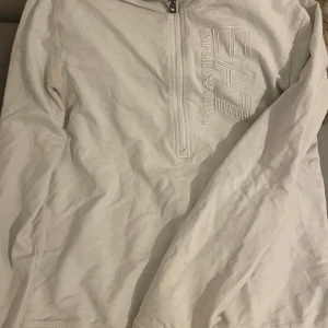 Vit EA7 långärmad tröja med half zip - Säljer en stilren vit långärmad tröja från EA7 Emporio Armani med half zip framtill. Tröjan har ett stort, upphöjt EA7-logotryck på bröstet och är gjord i mjuk bomull. Perfekt för dig som gillar clean och sportig look.