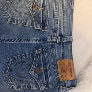 True Religion blå jeans bootcut - Säljer ett par klassiska blå jeans från True Religion med ikoniska bakfickor och snygga kontrastsömmar. Jeansen har bootcut-modell och är tillverkade i mjukt denim med detaljerade knappar och läderpatch bak. Perfekta för dig som gillar streetwear och vill sticka ut.