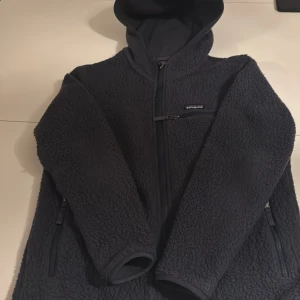 Mörkblå fleece från Patagonia storlek M - Mysig mörkblå fleece från Patagonia med huva och hel dragkedja. Jackan har två sidofickor med dragkedja och en bröstficka med dragkedja. Perfekt för kalla dagar och riktigt skön att bära. Klassisk Patagonia-logga på bröstet. Storlek M