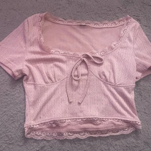 Ljusrosa croppad blus med spets - Superfin ljusrosa croppad blus med korta ärmar, spetsdetaljer vid halsringning och nederkant samt en söt knytning framtill. Blusen har ett diskret mönster i tyget och ger en romantisk vibe. Perfekt för dig som gillar gulliga och feminina toppar. Köpt i usa