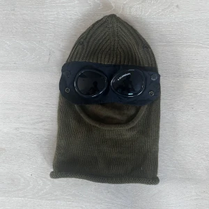 Olivgrön C.P. Company goggle-mössa - Säljer en olivgrön balaclava från C.P. Company med ikoniska svarta goggle-glasögon framtill. Mössan är stickad i mjukt material och har en cool, urban look med rundad öppning för ansiktet. Perfekt för dig som vill sticka ut med streetwear-vibe.