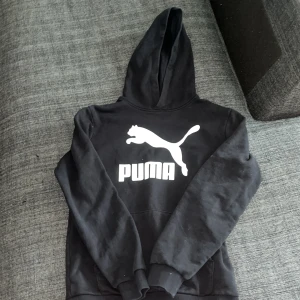 Svart hoodie från Puma med logga - Svart hoodie från Puma med stor vit logga och text på bröstet. Klassisk modell med huva och ribbade muddar. Perfekt för en sportig och chill stil. Tillverkad i mjuk bomullsblandning som är skön att ha på sig.
