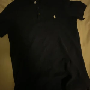 Mörkblå pikétröja från Ralph Lauren - Snygg mörkblå pikétröja från Polo Ralph Lauren med klassisk krage, knappar och den ikoniska broderade loggan på bröstet. Tröjan är kortärmad och gjord i bomull, perfekt för en clean och stilren look.