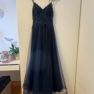 Mörkblå långklänning med spets och paljetter - Säljer en elegant mörkblå långklänning med tunna axelband. Klänningen har v-ringning och överdelen är dekorerad med blommig spets och glittrande paljetter. Kjolen är i flera lager tunn tyll som ger ett svepande intryck. Perfekt för bal eller fest!