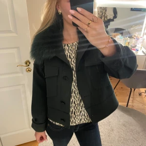 Meotine jacka - Snygg mörkgrön jacka från Meotine med stor fluffig pälskrage. Jackan har två stora fickor framtill, knappar och en croppad passform. Perfekt för dig som vill sticka ut med en cool och trendig look. Materialet är ull och päls, vilket ger en lyxig känsla.