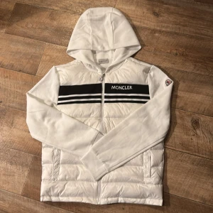 Vit Moncler cardigan med huva - Snygg vit pufferjacka från Moncler med huva och svarta ränder över bröstet. Jackan har dragkedja framtill, långärmade ärmar och broderad Moncler-logga på axeln. Materialet är dun och polyester, perfekt för dig som gillar stilren design med sportig touch. Storlek m men passar s också