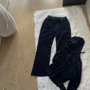 Juicy Couture mörkblå velour set - Mörkblå velour sett från Juicy Couture med dragkedja framtill, huva med snörning och broderad JC-logga på bröstet. Västen har en klassisk hoodie-look och är supermjuk med känguruficka framtill. Perfekt för en chill och trendig stil. Säljs även styck vis men billigare sett