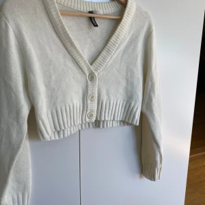Vit kofta ifrån H&M - Säljer en vit  kofta från H&M med ribbade muddar och v-ringad hals. Koftan har tre knappar framtill och är stickad i mjukt material. Perfekt för lager-på-lager och chill outfits. Långärmad och med snygg passform som ger en trendig vibe.