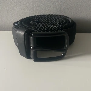 Svart flätat bälte 80 cm - Snyggt svart flätat bälte med klassiskt spänne i svart metall. Bältet har detaljer i svart skinn vid ändarna och är gjort i ett flexibelt, vävt material som ger en cool och modern look. Perfekt för att lyfta vilken outfit som helst.