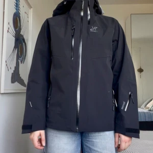 Svart skaljacka RevolutionRace M - Svart skaljacka från RevolutionRace med huva och flera dragkedjeförsedda fickor. Jackan är av modellen hypershell pro och har där av vattentålig och vindtålig. Därav är den perfekt för skidåkning eller andra outdoor aktiviteter. 