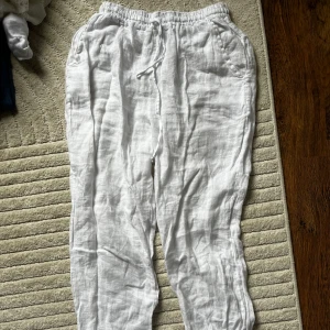 Vita linnebyxor från Design by si - Snygga vita linnebyxor från Design by Si med avslappnad passform och elastisk midja med snörning. Byxorna har fickor på sidorna och är gjorda i mjukt linne, perfekta för varma dagar. 